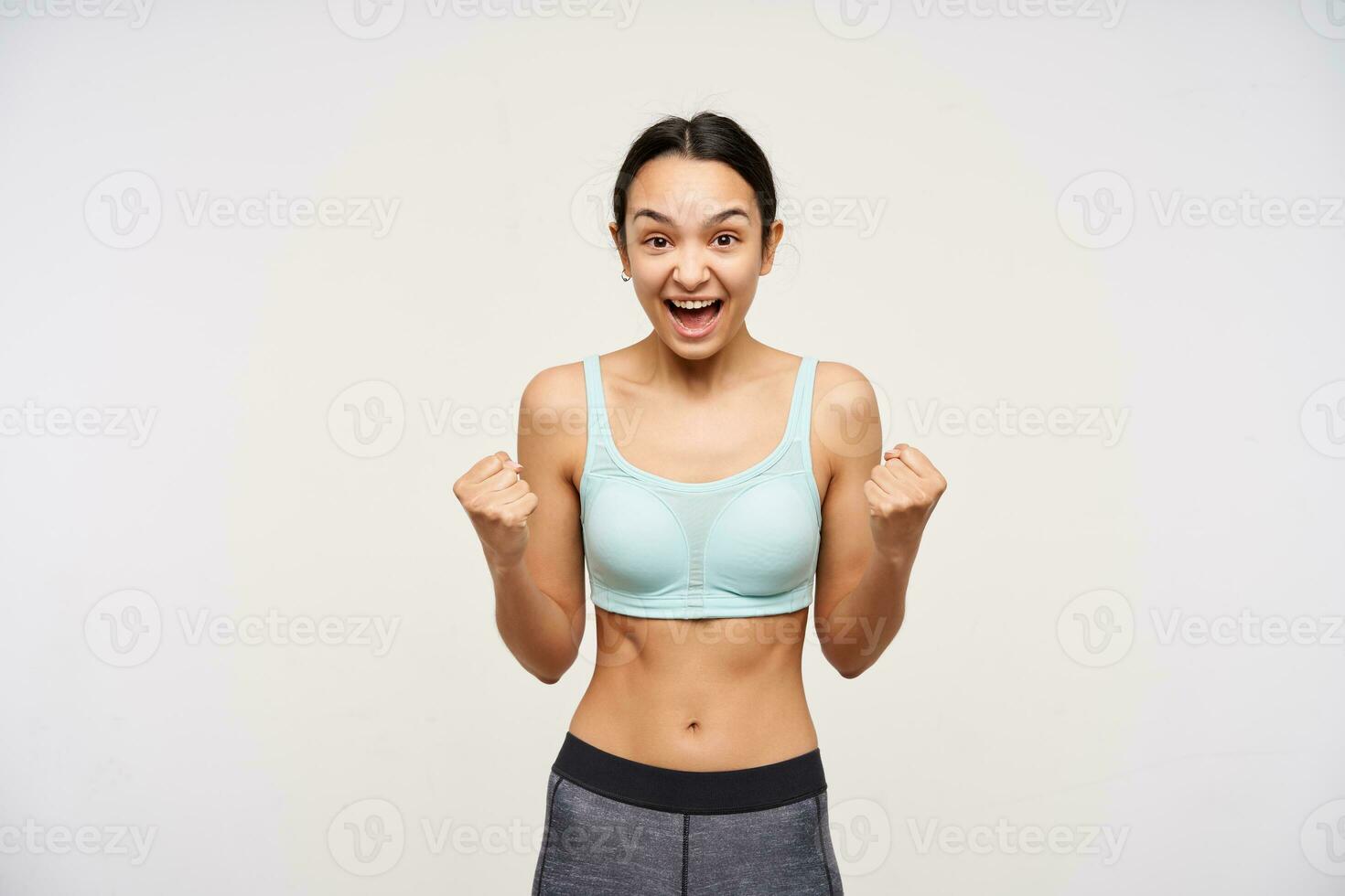 Energized young brown haired slim female raising emotionally her fists while rejoicing about something with wide mouth opened, isolated over white background photo