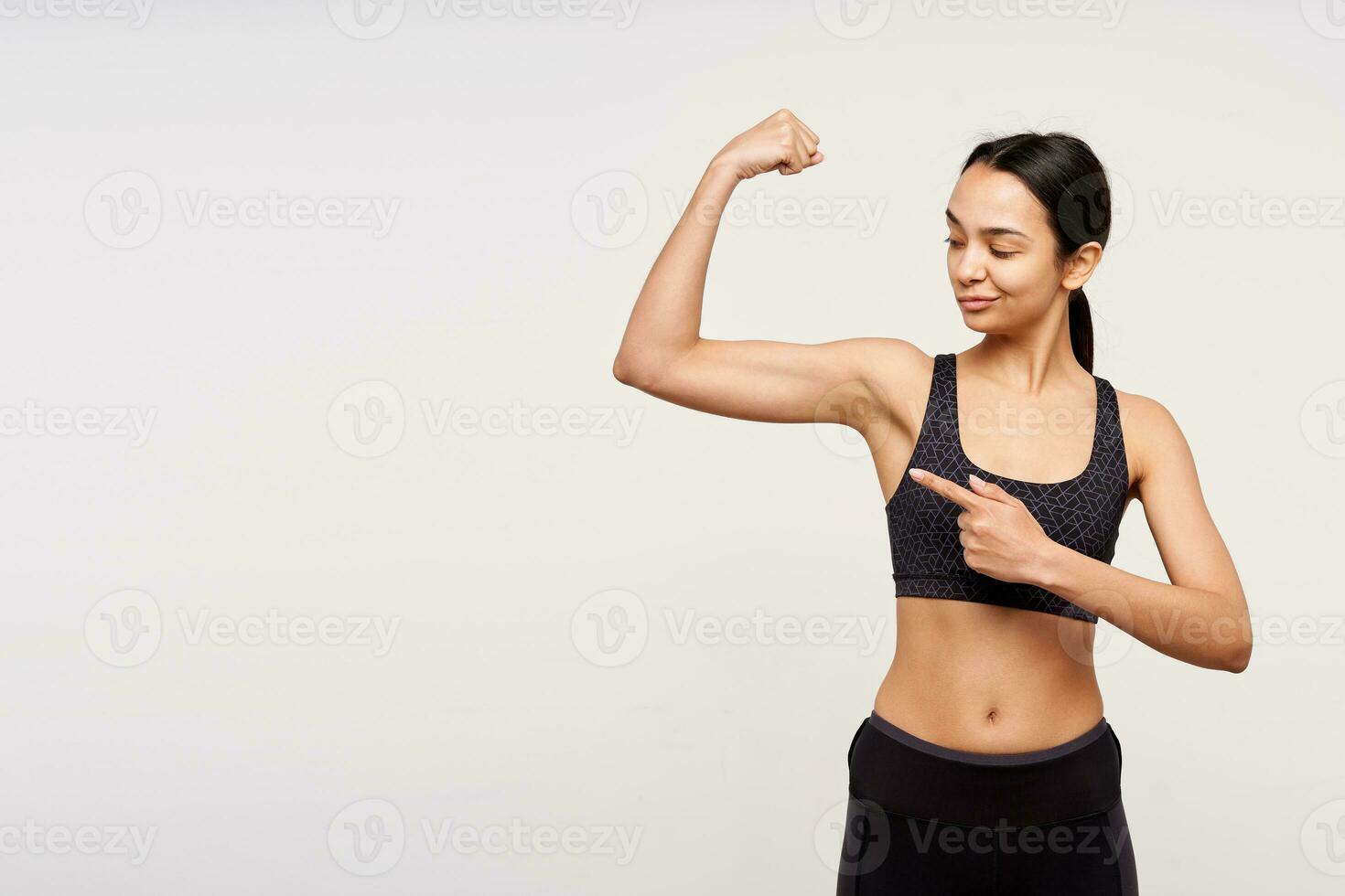 Confident young pretty dark haired woman in sporty wear twisting her mouth while looking on her strong hand and showing on it with forefinger, posing over white background photo