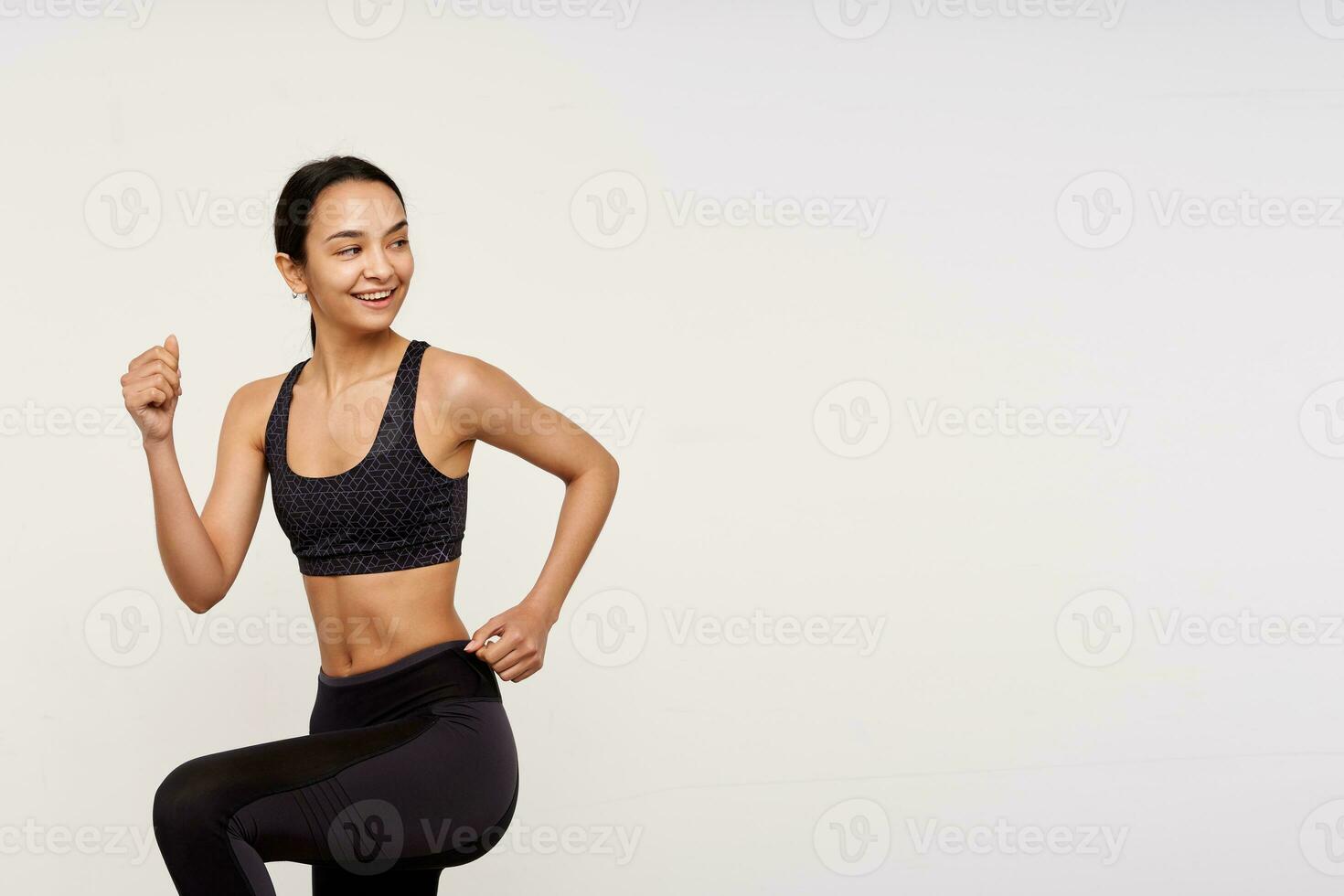 Studio shot of glad slim young brown haired female lady with casual hairstyle being in nice mood while making morning workout, standing over white background photo