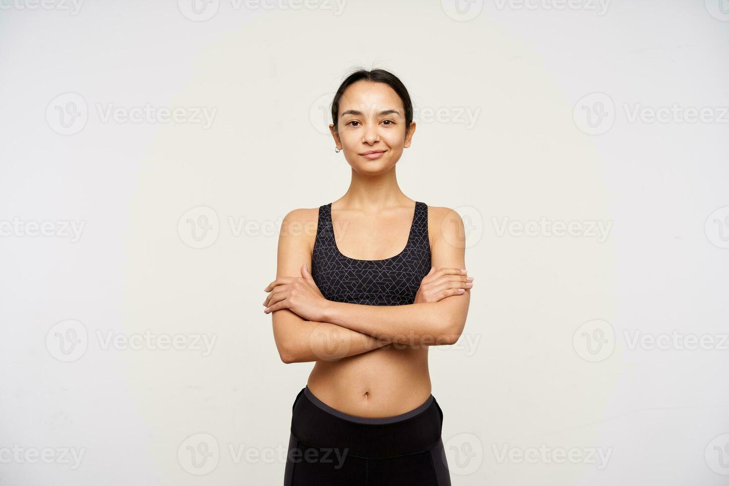 estudio foto de joven Delgado marrón peludo mujer en deportivo vestir plegable su manos en cofre mientras mirando afirmativamente a cámara con amable sonrisa, en pie terminado blanco antecedentes