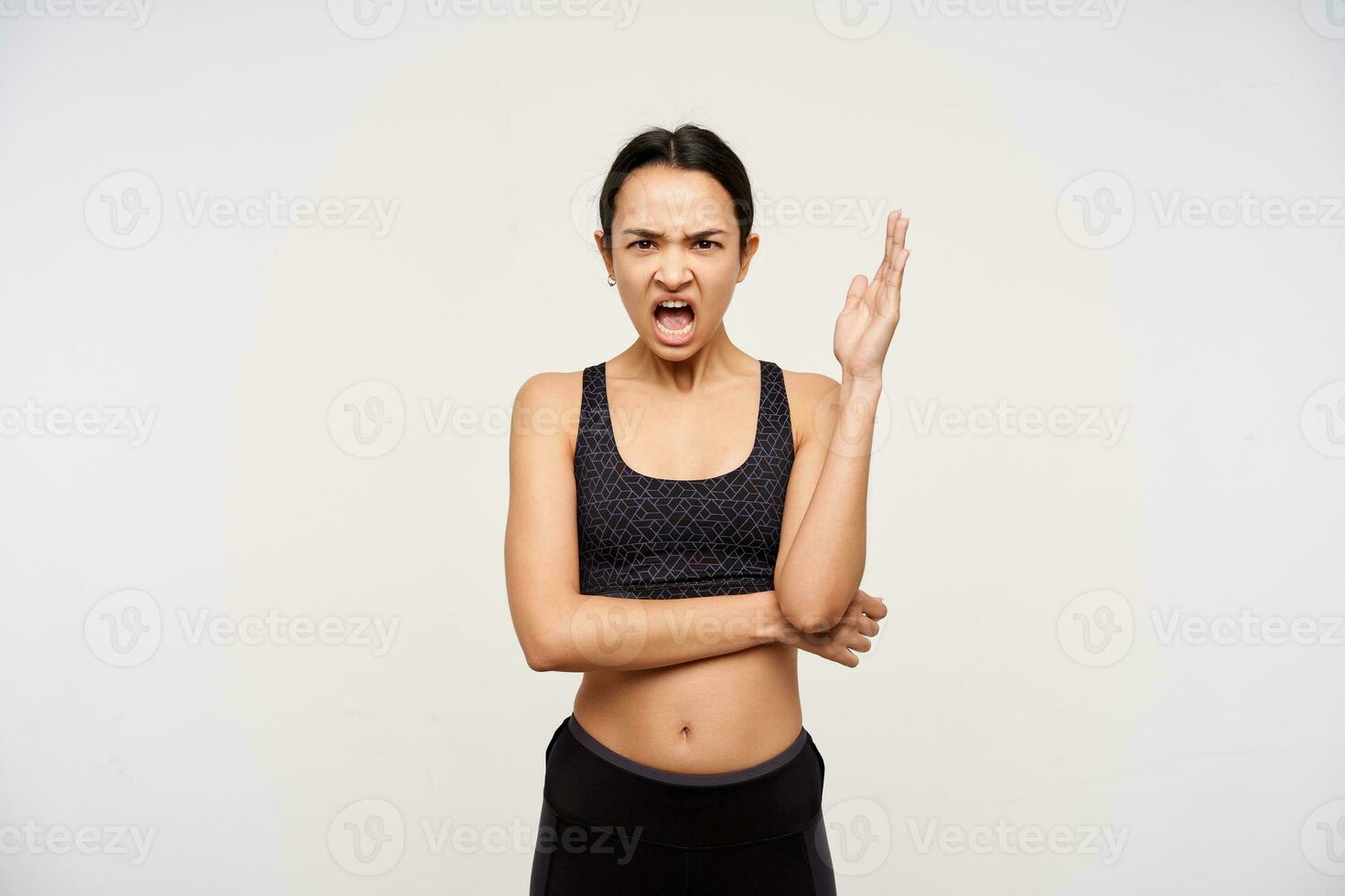 Indoor photo of young brunette woman with casual hairstyle keeping her palm raised and frowning eyebrows while looking angrily at camera, isolated over white background