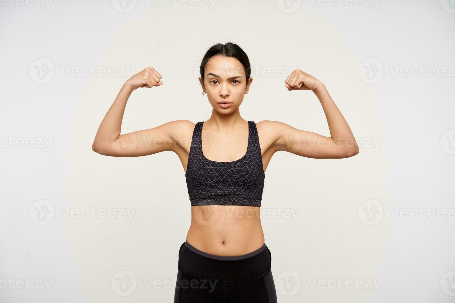 Severe young sporty brown haired woman raising her hands while demonstrating strong biceps and looking seriously at camera, isolated over white background photo