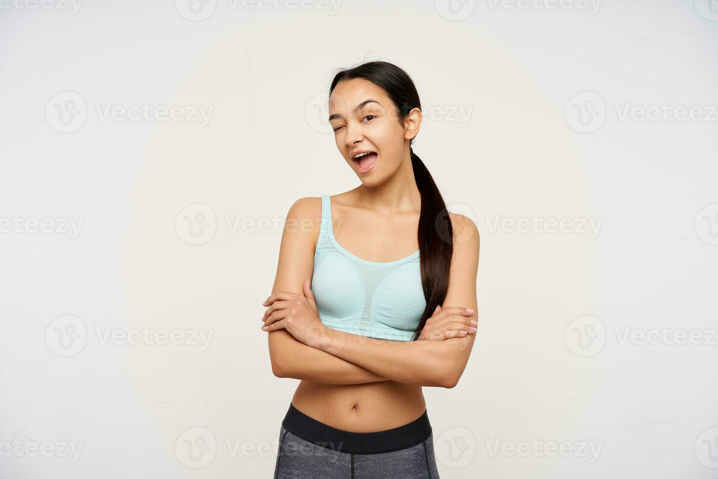 Portrait of attractive, adult asian girl with dark long hair. Wearing sportswear and flirty winking to you with arms crossed on a chest. Watching at the camera isolated over white background photo