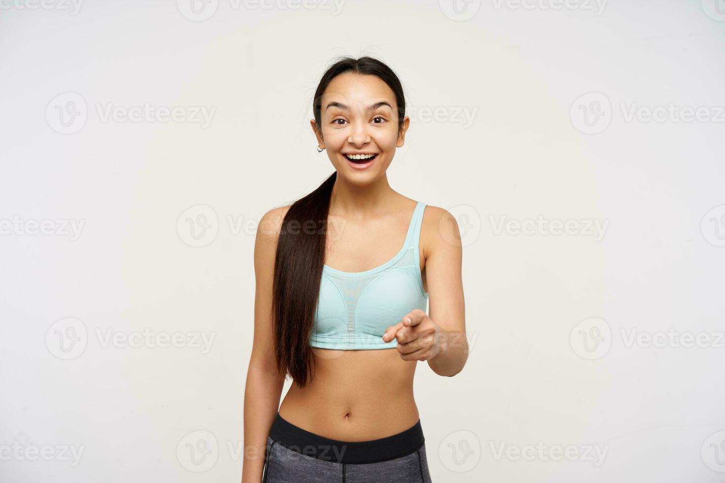 Young lady, sporty, pretty asian woman with dark long hair gathered in a ponytail. Wearing sportswear and pointing with big smile at you. Watching at the camera isolated over white background photo