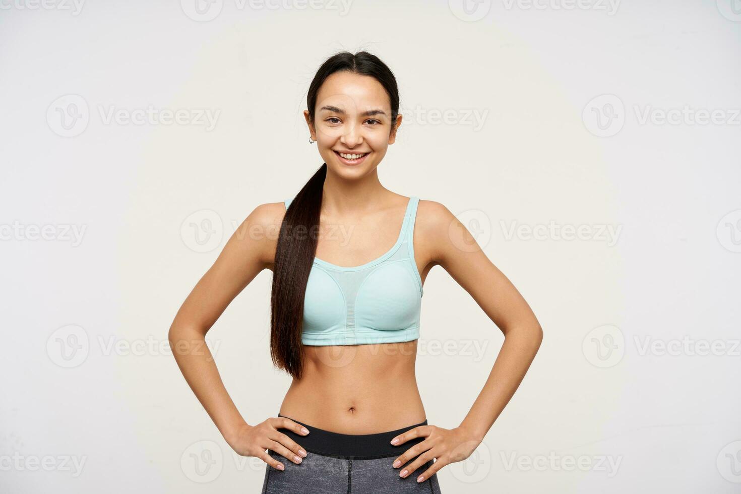 Young lady, sporty asian woman with dark long hair gathered in a ponytail. Wearing sportswear and holding hands on a waist, smiling. Watching at the camera isolated over white background photo