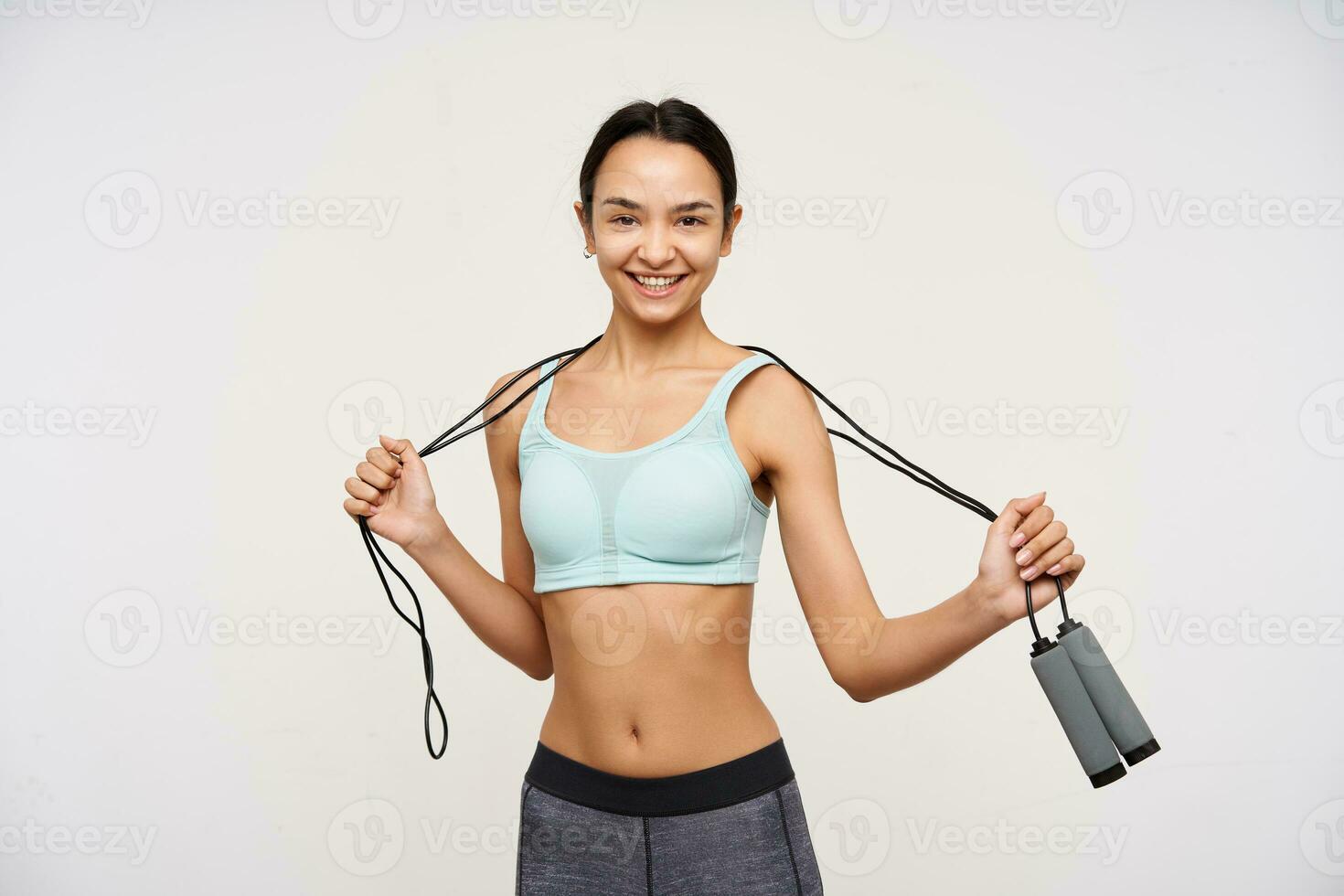 Portrait of attractive, sporty asian girl with dark long hair. Wearing sportswear and holding a jump rope over her neck. Have a joyful smile. Watching at the camera isolated over white background photo