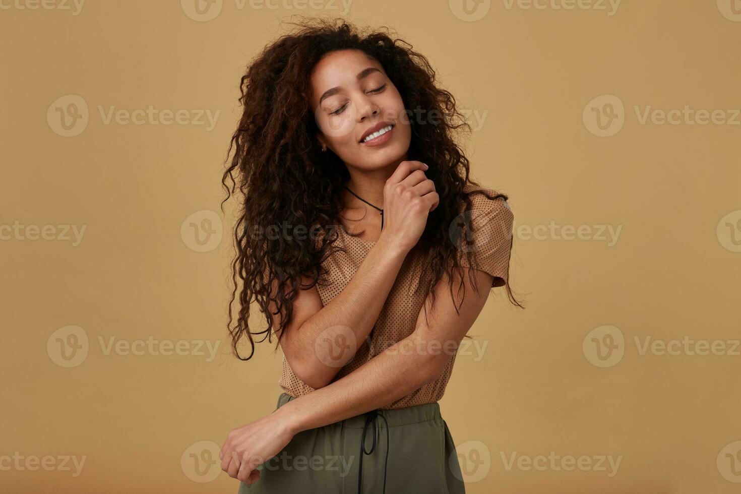 Goog looking young glad brown haired curly dark skinned woman keeping her eyes closed while smiling pleasantly and embracing her with raised hands, isolated over beige background photo