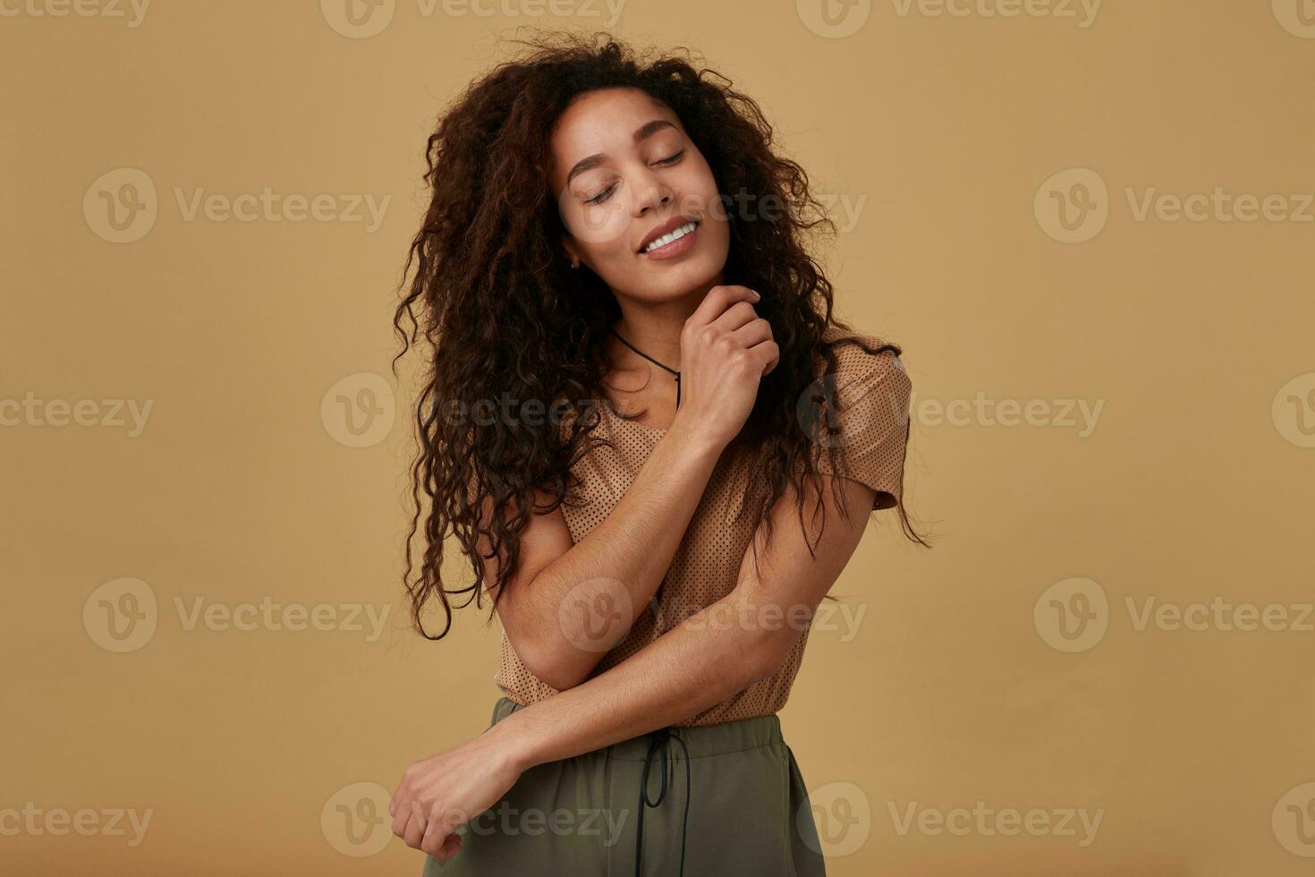 Goog looking young glad brown haired curly dark skinned woman keeping her eyes closed while smiling pleasantly and embracing her with raised hands, isolated over beige background photo