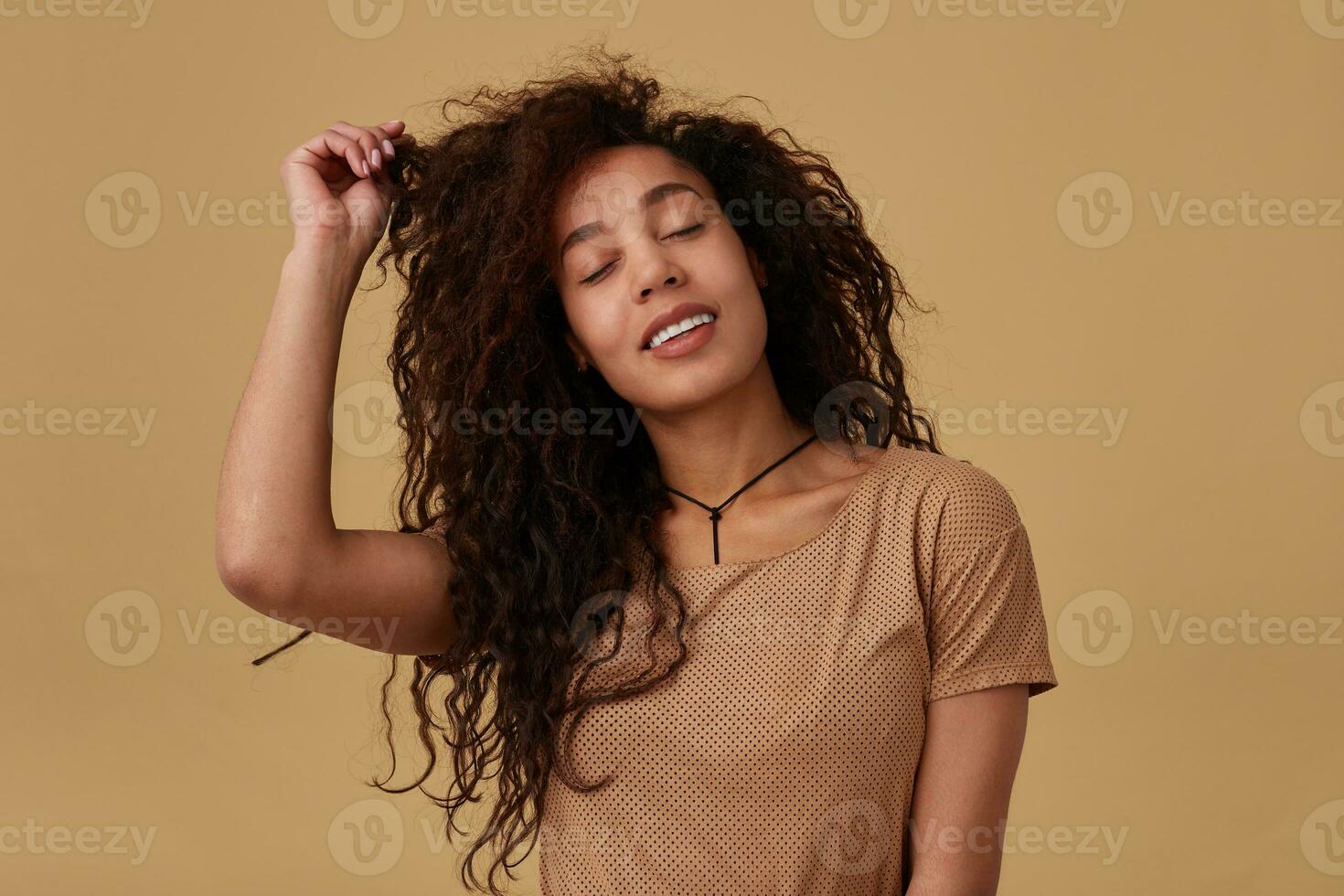 Pleasant looking young brown curly lady with dark skin touching her hair with raised hand and smiling gladly with closed eyes, isolated over beige background photo