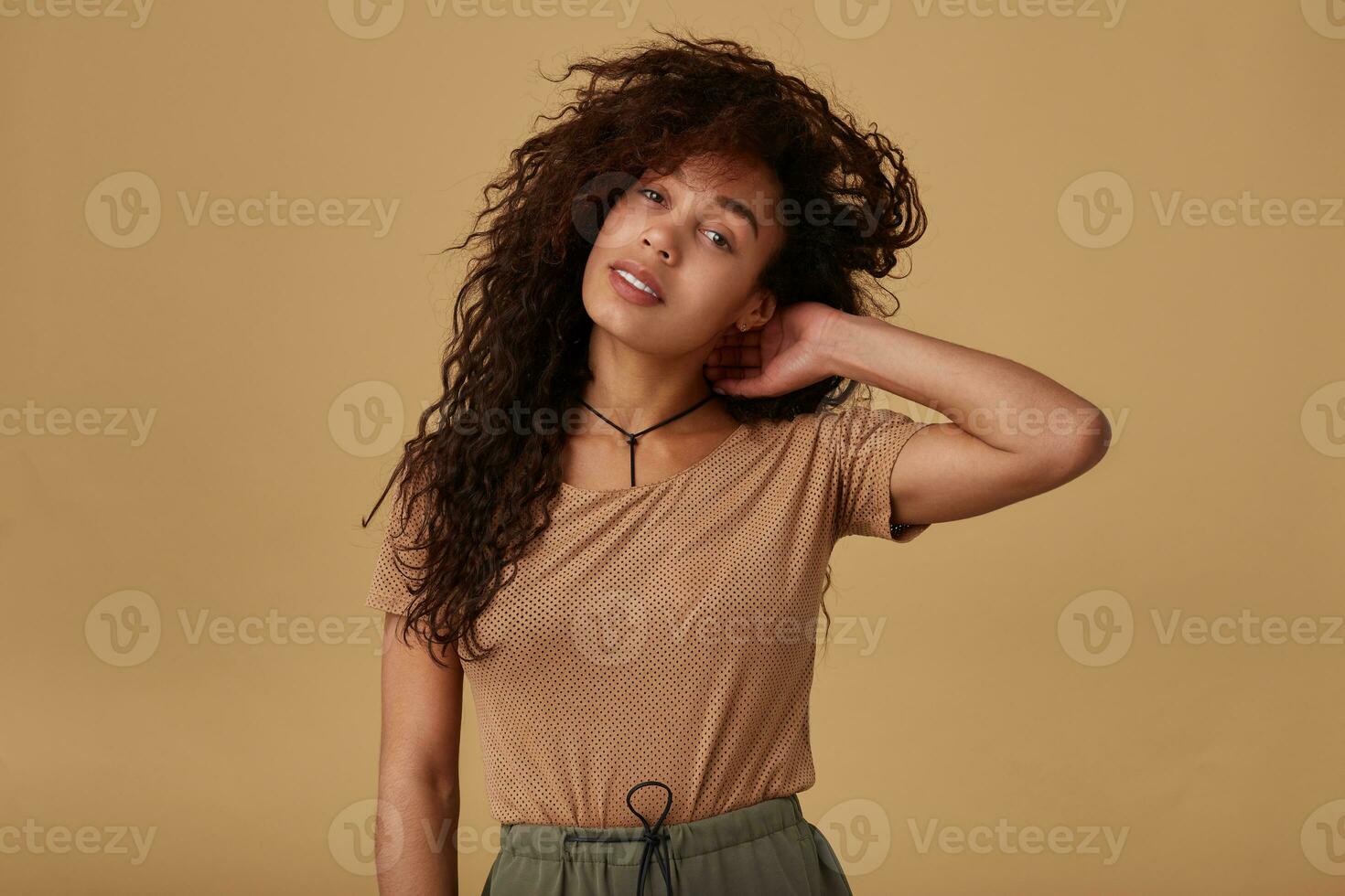 Attractive young dark skinned brunette woman with wild curly hair raising hand to her head and looking calmly at camera, standing over beige background photo