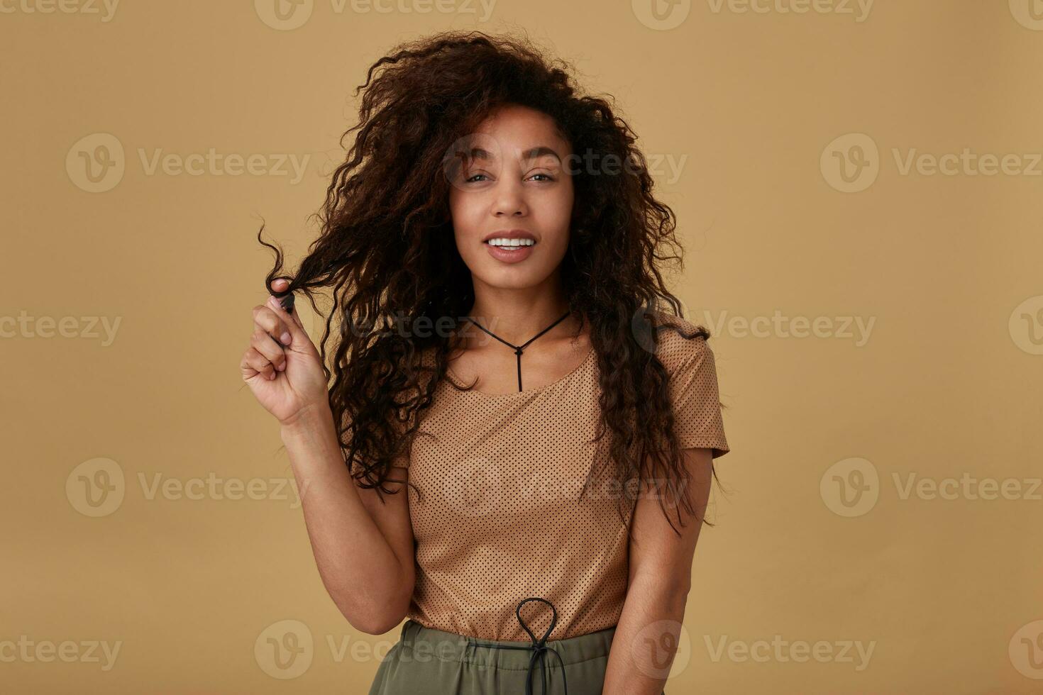 Cheerful young pretty dark skinned lady twisting her brown curly hair on raised finger and smiling gladly at camera while posing over beige background in casual wear photo