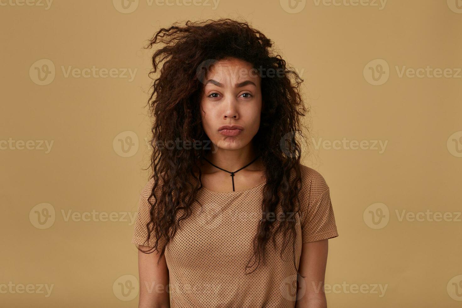 Bewildered young brown-eyed curly brunette woman with dark skin frowning confusedly her eyebrows while looking at camera, isolated over beige background photo