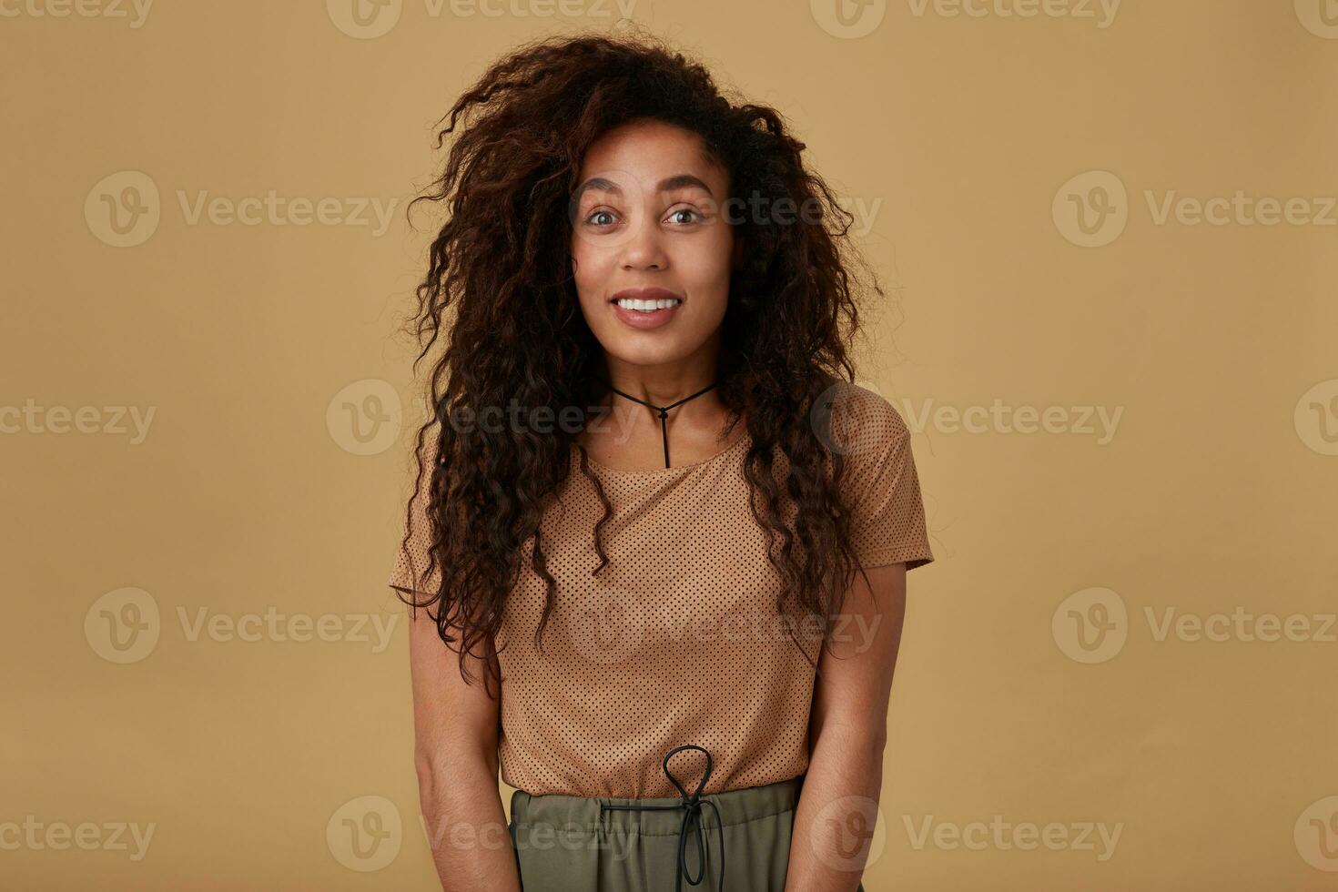 Glad young pretty long haired curly dark skinned lady with brown loose hair looking joyfuuly at camera and keeping hands down while posing over beige background photo