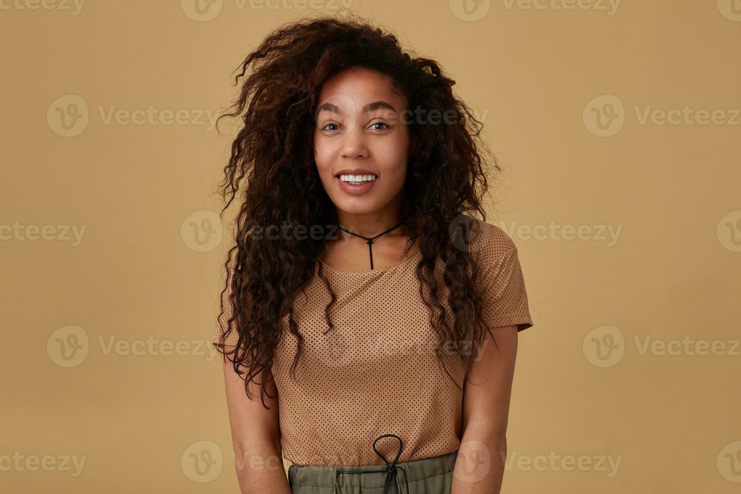 Studio shot of young lovely brown haired curly woman with dark skin looking cheerfully at camera with wide happy smile while standing over beige background photo