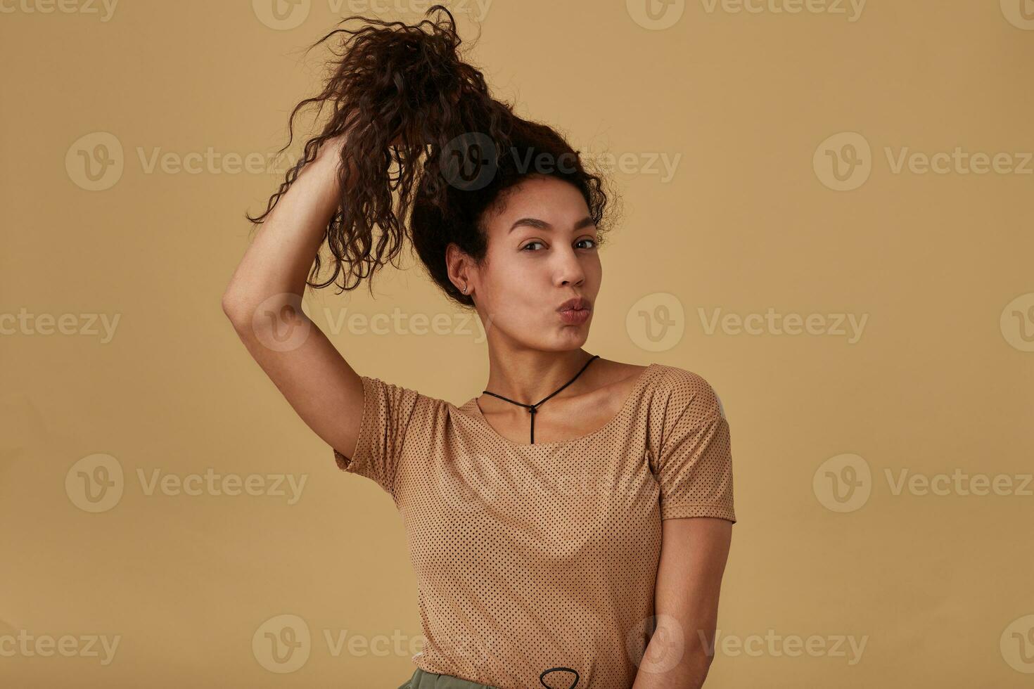 Funny young pretty dark skinned brunette woman keeping her curly hair with raised hand and pursing lips while looking at camera, isolated over beige background photo