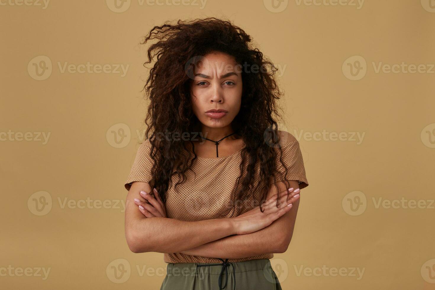 ofendido joven bonito Rizado oscuro desollado morena dama plegable su manos en cofre mientras mirando tristemente a cámara y ceñudo su Cejas, posando terminado beige antecedentes foto