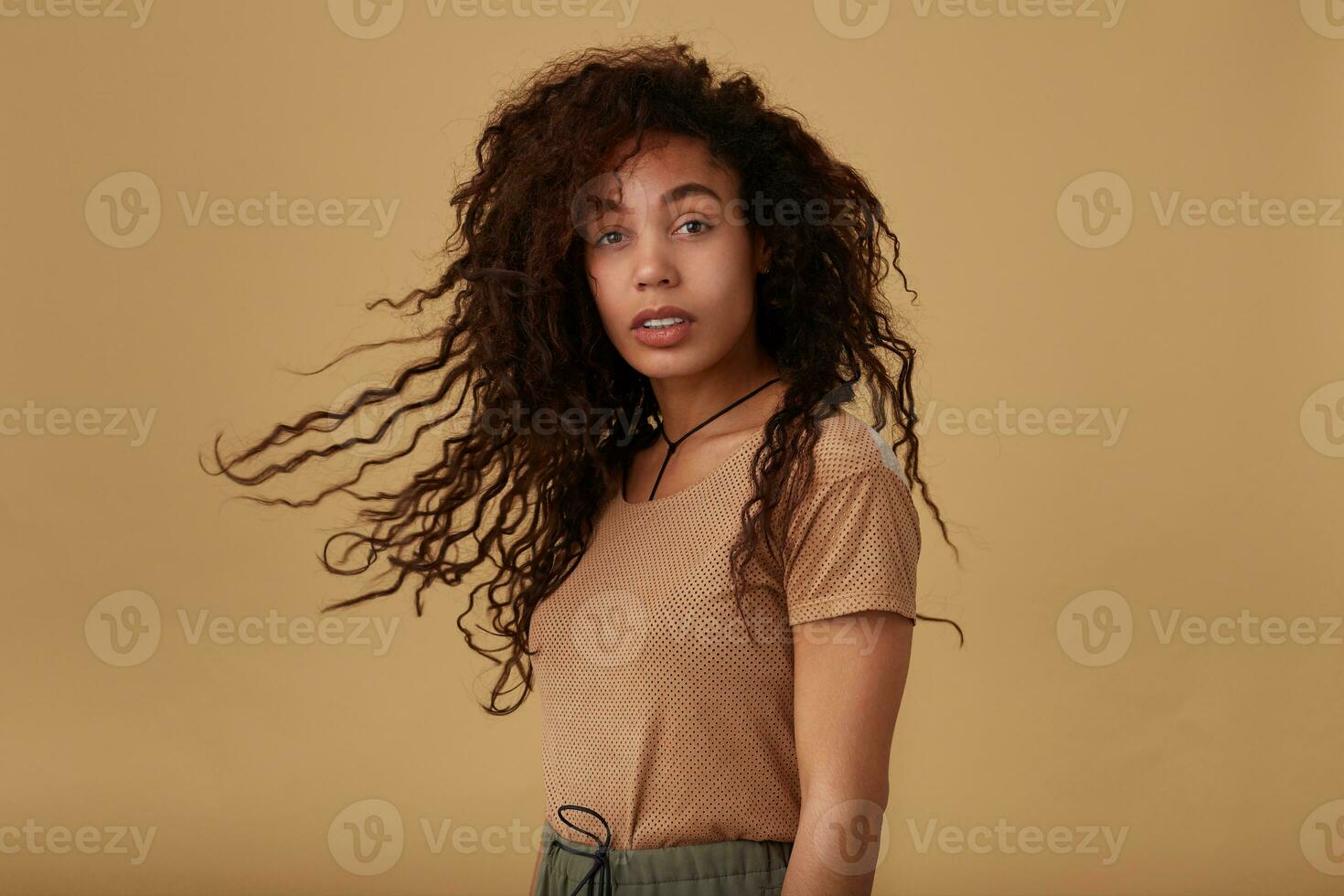 Studio shot of young dark skinned brunette female waving her long curly hair and looking calmly at camera while standing over beige background with hands down photo