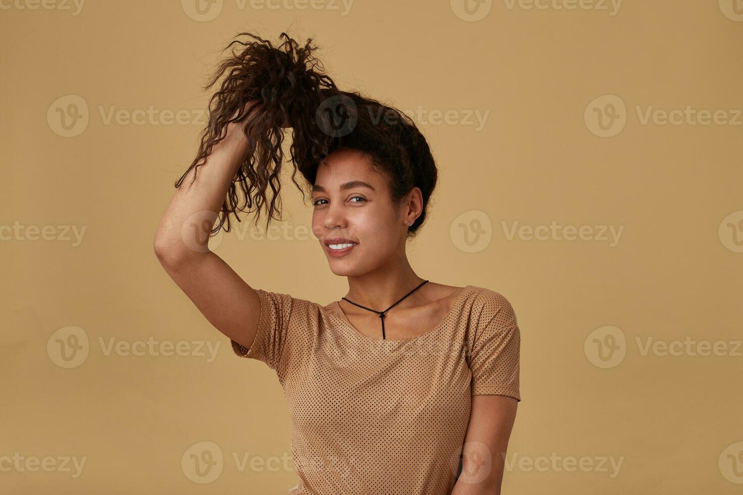 Positive young lovely brown haired curly female with dark pulling her hair with raised hand and smiling cheerfully at camera, isolated over beige background photo