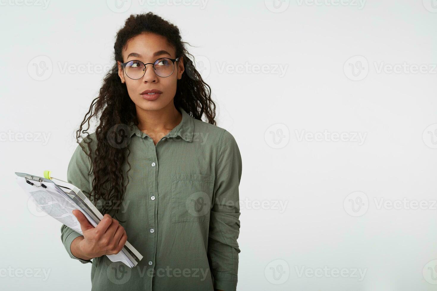 interior Disparo de joven atractivo Rizado morena oscuro desollado hembra mirando pensativamente hacia arriba mientras en pie terminado blanco antecedentes con libros de texto en su manos foto