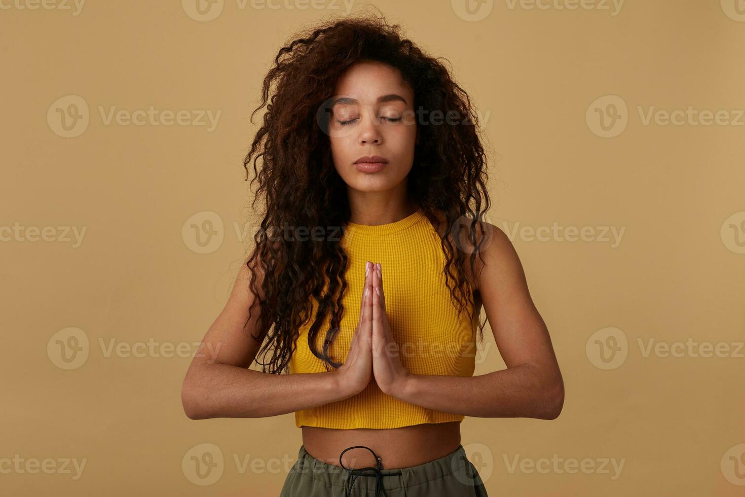 Pleasant looking young calm curly brunette dark skinned woman keeping her eyes closed while meditating and raising folded hands, isolated over beige background photo