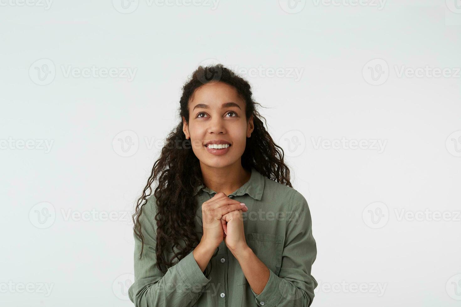 Indoor photo of cheerful brown haired curly dark skinned woman with casual hairstyle smiling widely while looking upwards and keeping raised hand together, isolated over white background
