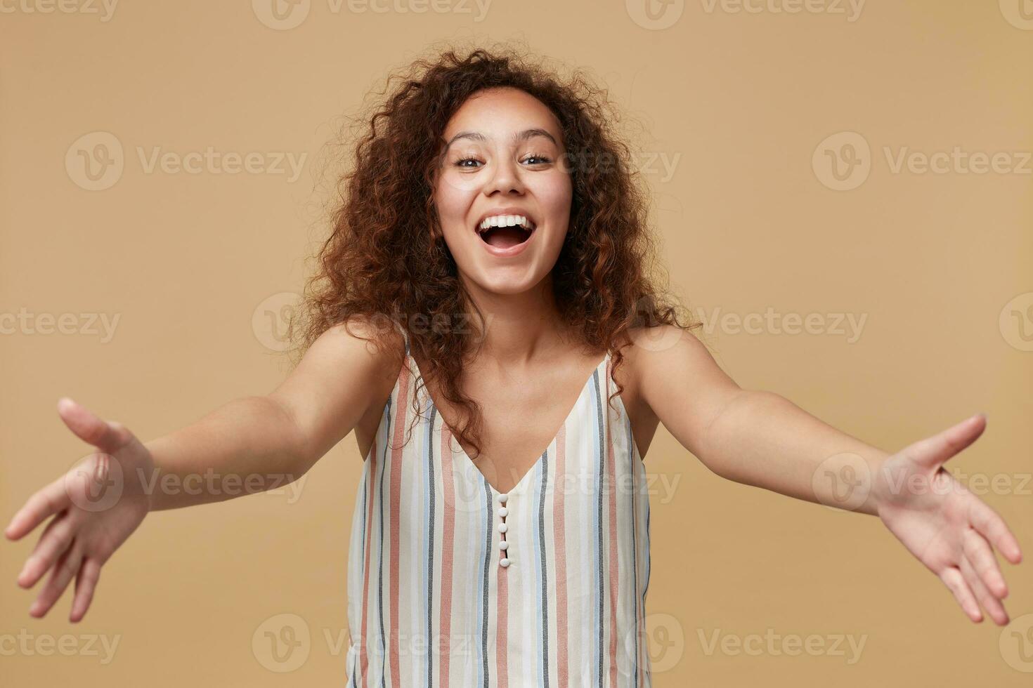 Portrait of young pretty cheerful brown haired curly female keeping her hands wide opened while looking joyfully at camera, isolated over beige background photo