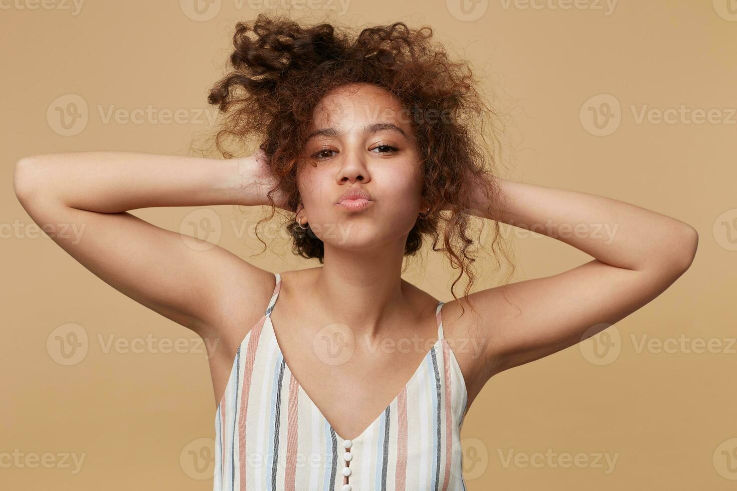 Portrait of positive young brown haired curly female keeping raised hands on her head and pursing lips while looking at camera, isolated over beige background photo