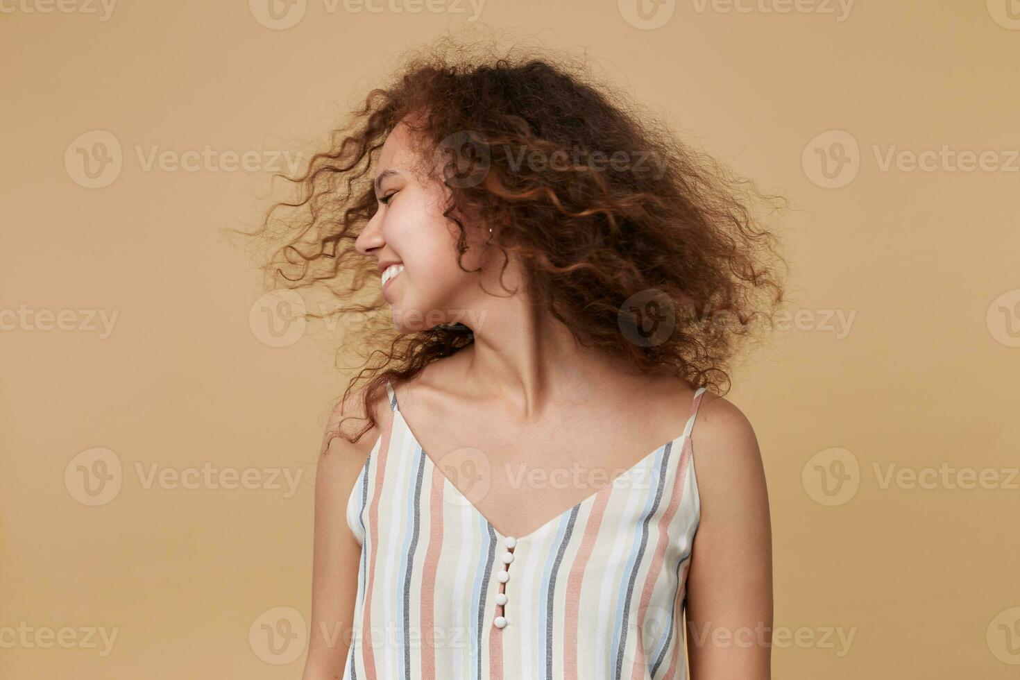 Good looking young pretty brown haired curly lady in summer top waving her head and smiling positively while standing over beige background with hands down photo