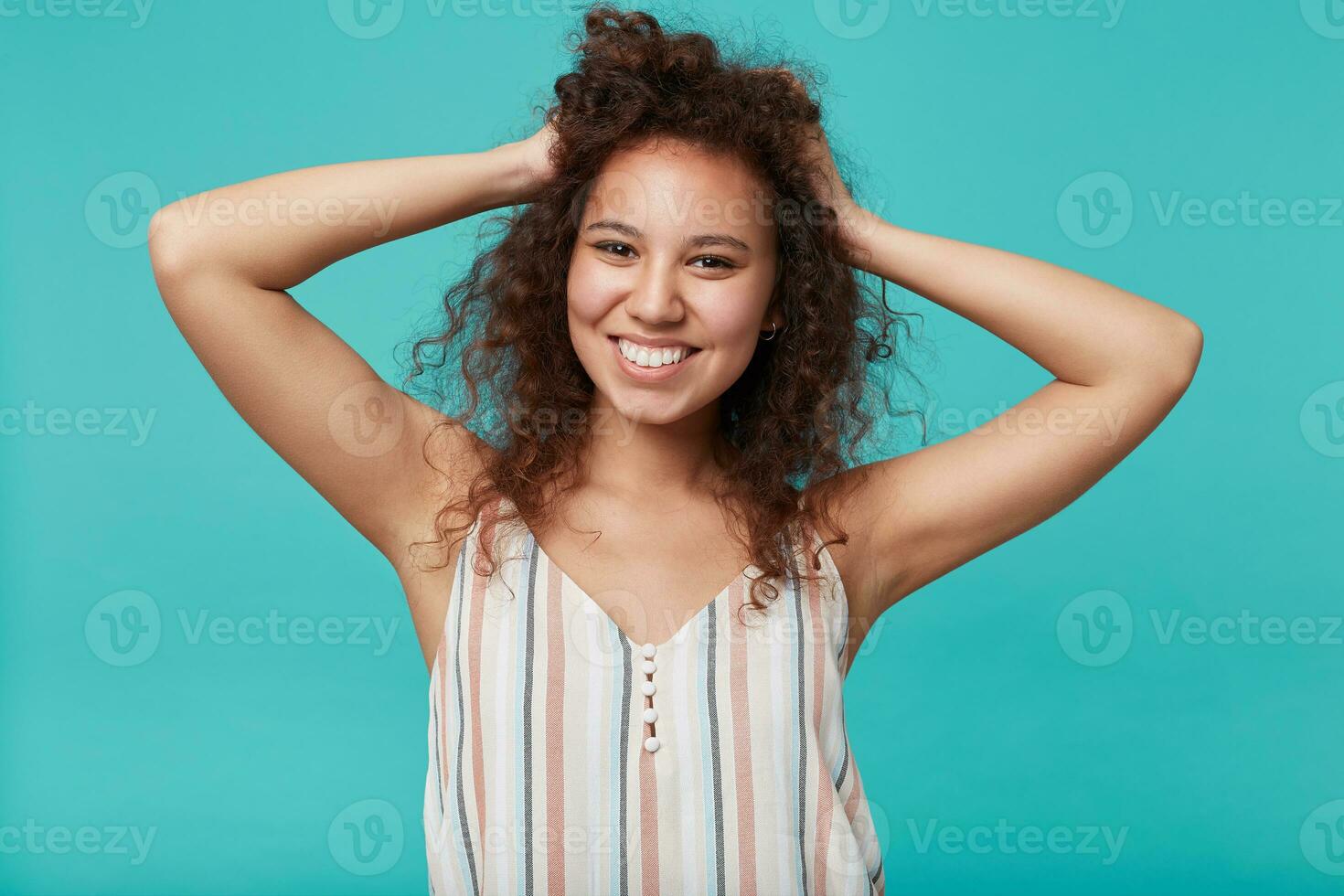 Studio photo of young attractive brown haired curly female looking at camera with charming smile and keeping raised hands on her head, isolated over blue background