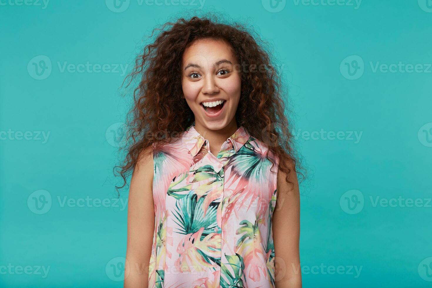 Portrait of young joyful brown-eyed long haired curly brunette female looking happily at camera with wide mouth opened while posing blue background with hands down photo