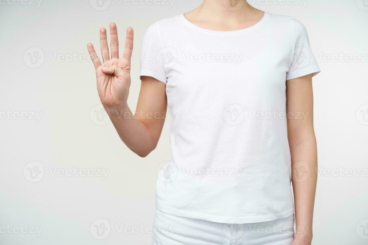 Studio photo of young fair-skinned female keeping hand raised while counting four with fingers, using sign language to talk to deaf person, isolated over white background