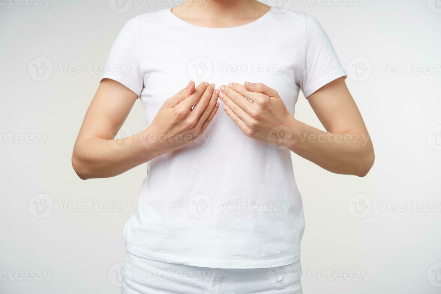 Studio photo of young female dressed in white t-shirt learning sign language while standing over white background, keeping palms in front of her chest while showing word book