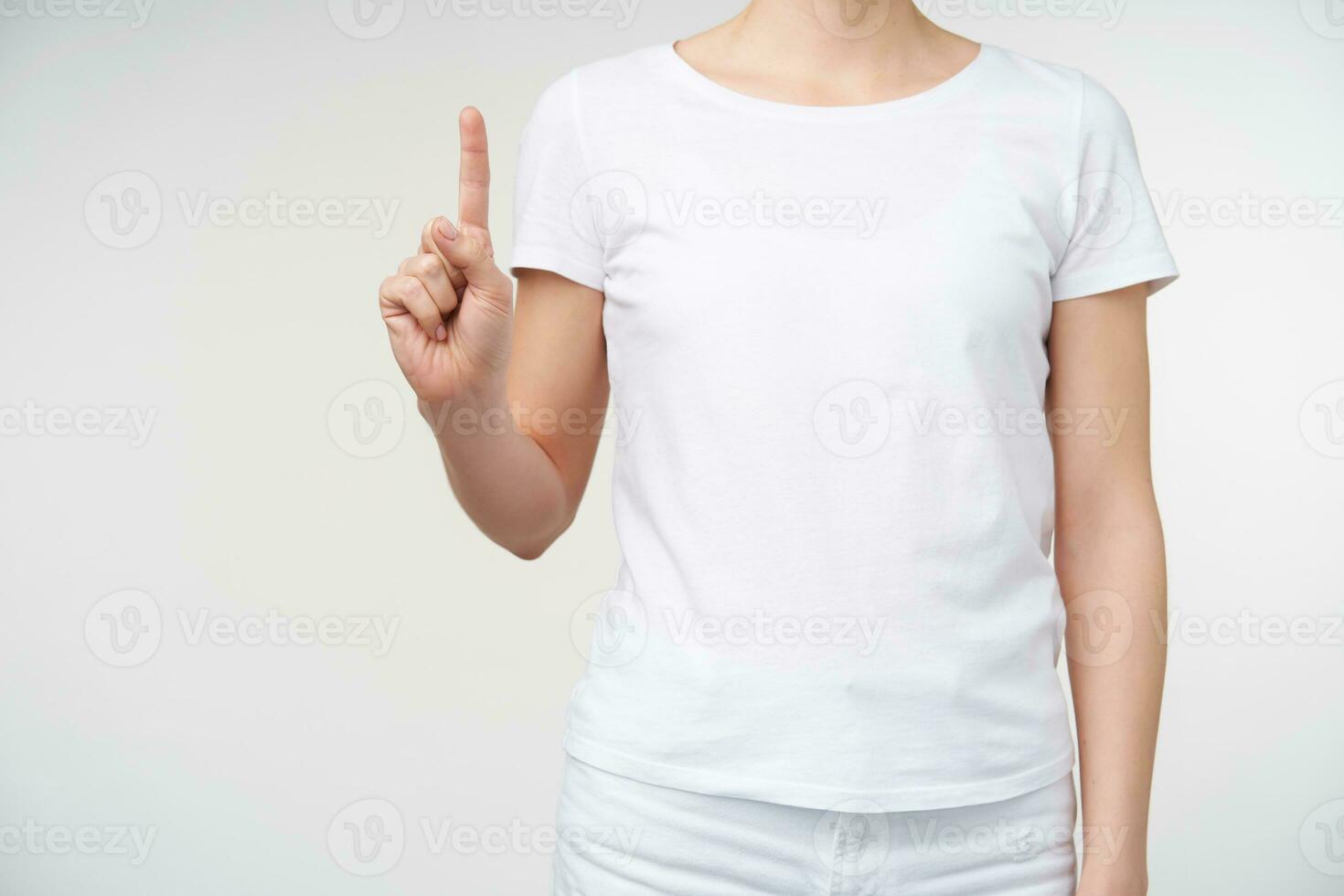 Studio photo of young woman dressed in white t-shirt while posing over white background, keeping forefinger raised while counting one. Hands and gesturing concept