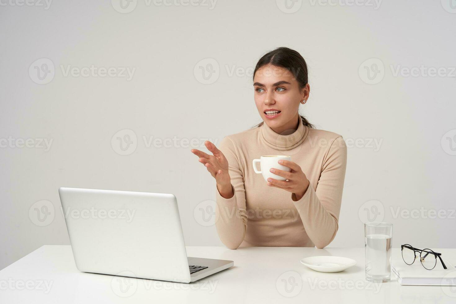 Beautiful young dark haired business lady having meeting with colleagues, holding cup of tea while sitting over white background in beige poloneck and keeping her hand raised photo