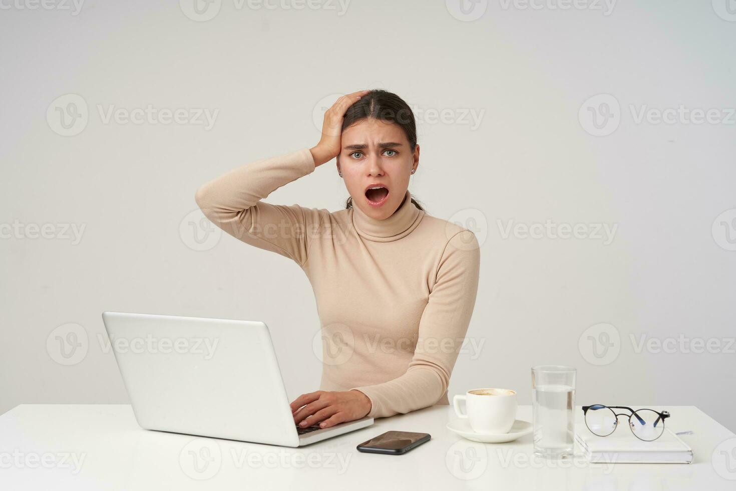 Amazed young blue-eyed brunette woman with elegant hairstyle keeping palm on her head while looking perplexedly at camera, frowning eyebrows with opened mouth, isolated over white background photo