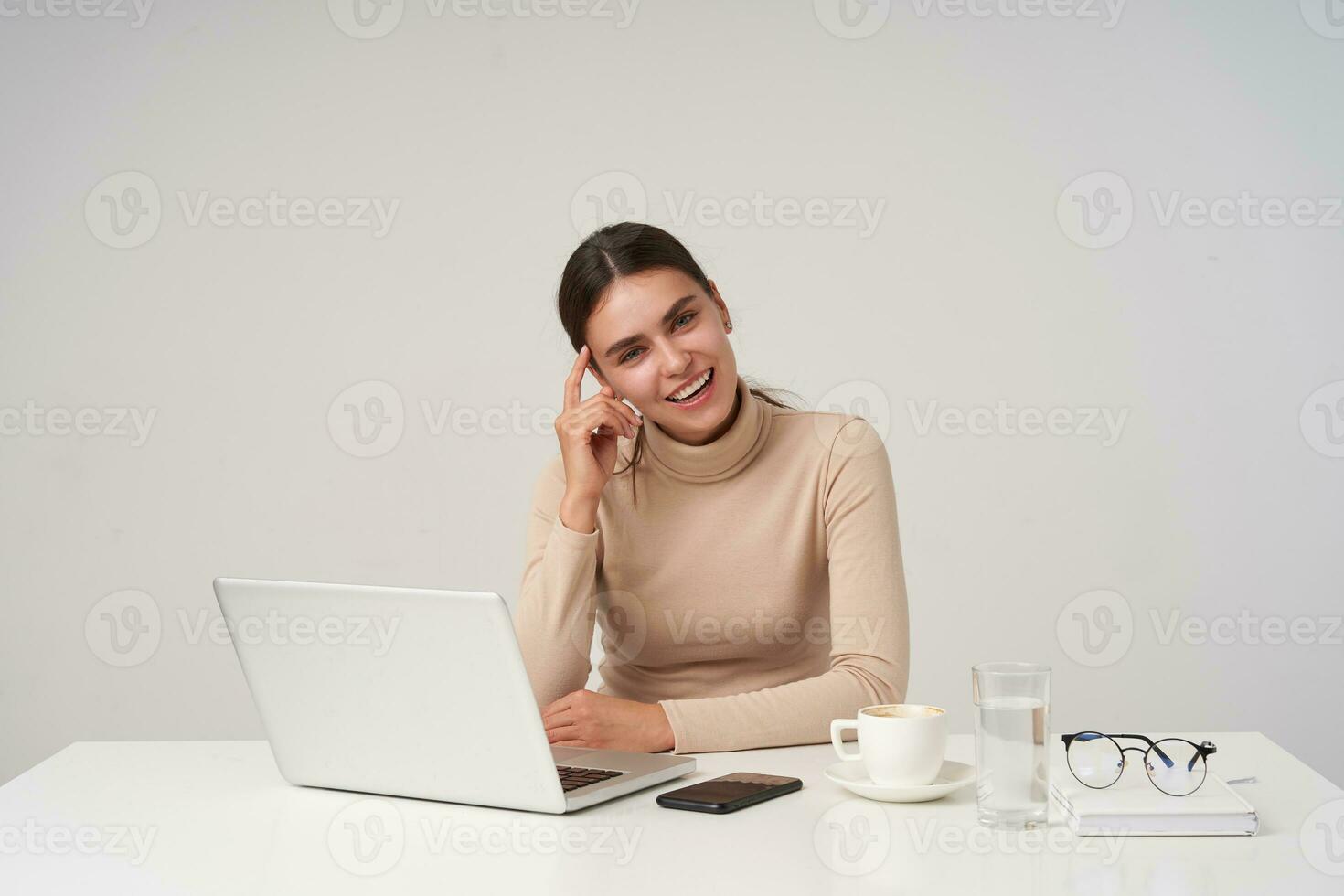 Positive young attractive brunette businesswoman with ponytail hairstyle touching her face with raised hand while sitting over white background and smiling cheerfully photo