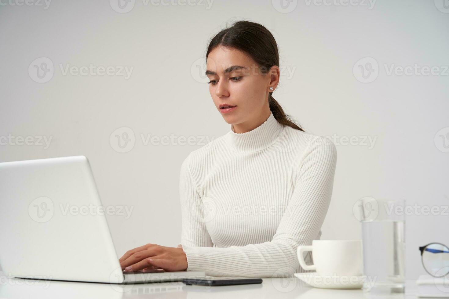 concentrado joven bonito morena mujer con natural maquillaje trabajando con su ordenador portátil y acuerdo manos en teclado, vestido en formal ropa mientras posando terminado blanco antecedentes foto