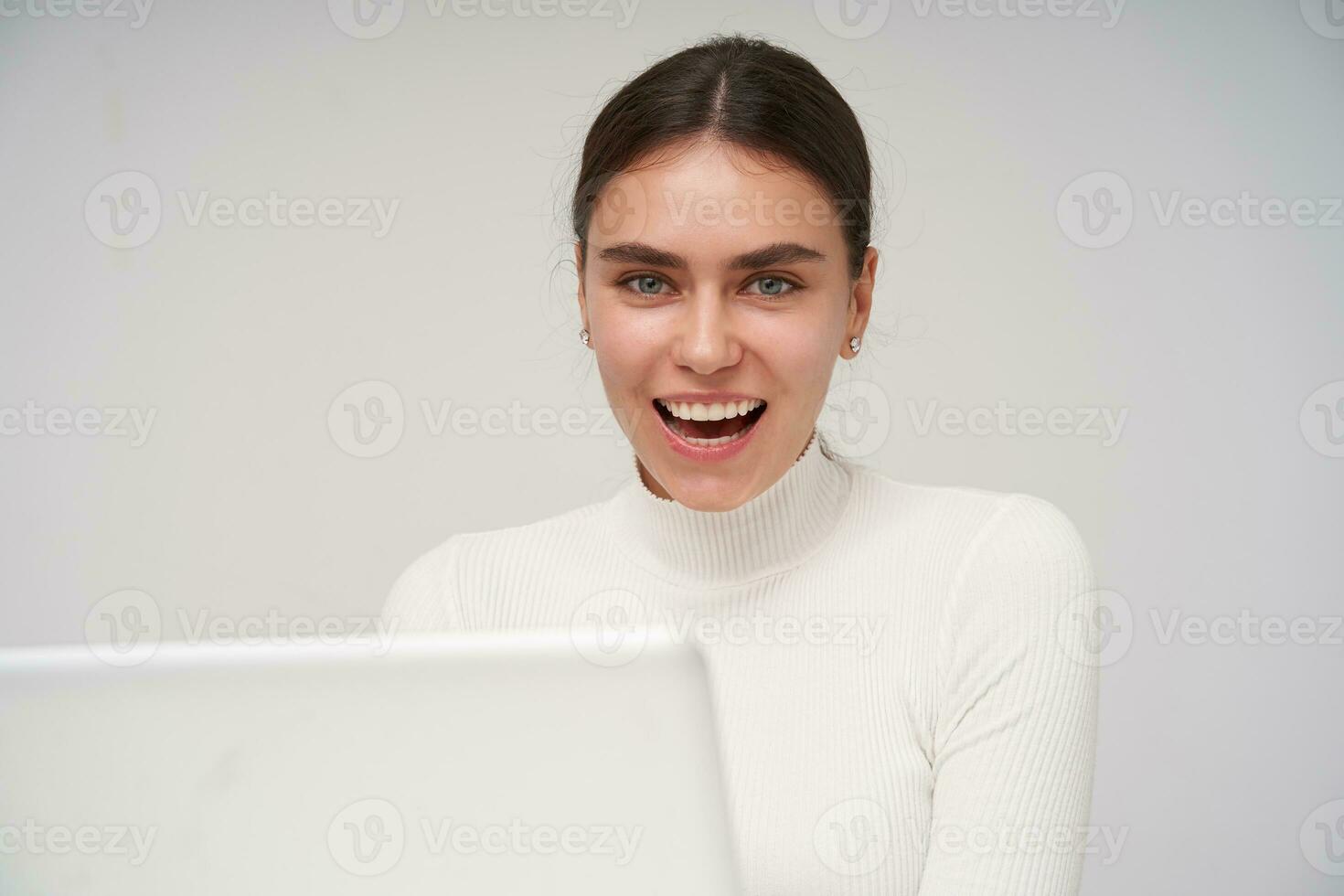 Portrait of young beautiful dark haired lady with ponytail hairstyle dressed in formal clothes being in high spirit while posing over white background, showing perfect white teeth while smiling widely photo