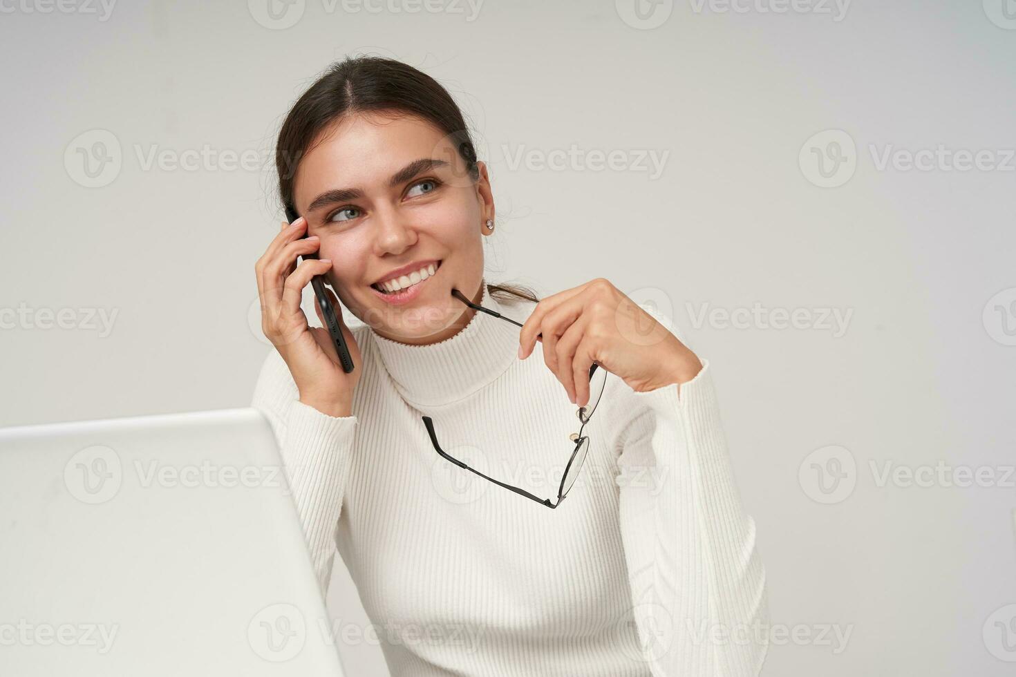 Closeup of young pretty brunette woman in white knitted poloneck smiling cheerfully while having phone converstation and holding glasses, isolated over white background photo