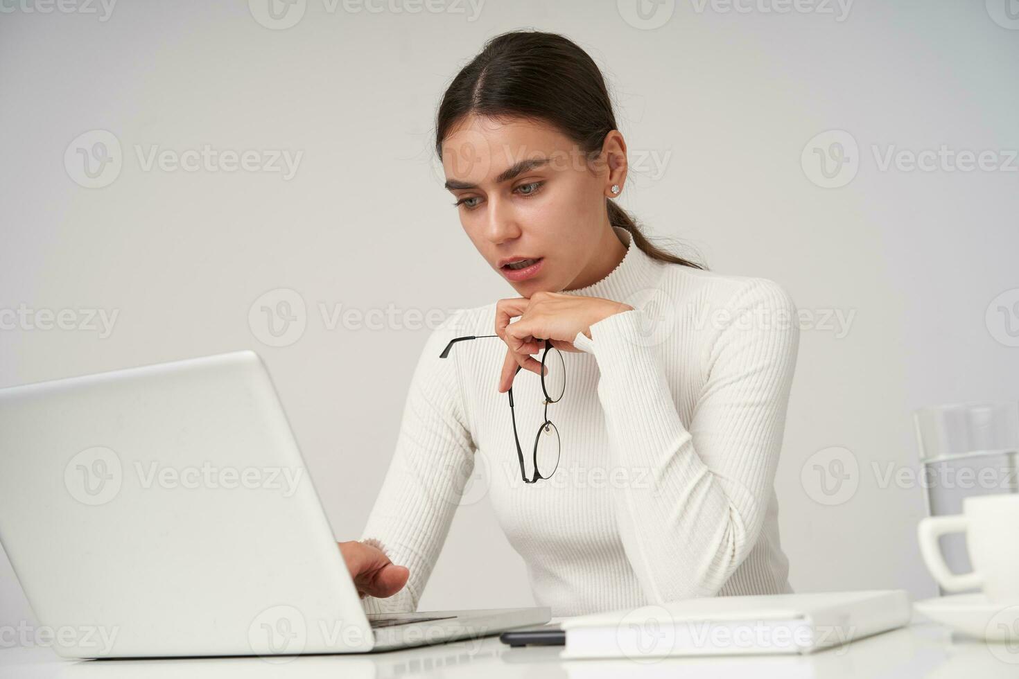 Concentrated young attractive brunette woman with natural makeup looking at screen of her laptop with serious face, dressed in formal clothes while sitting over white background photo