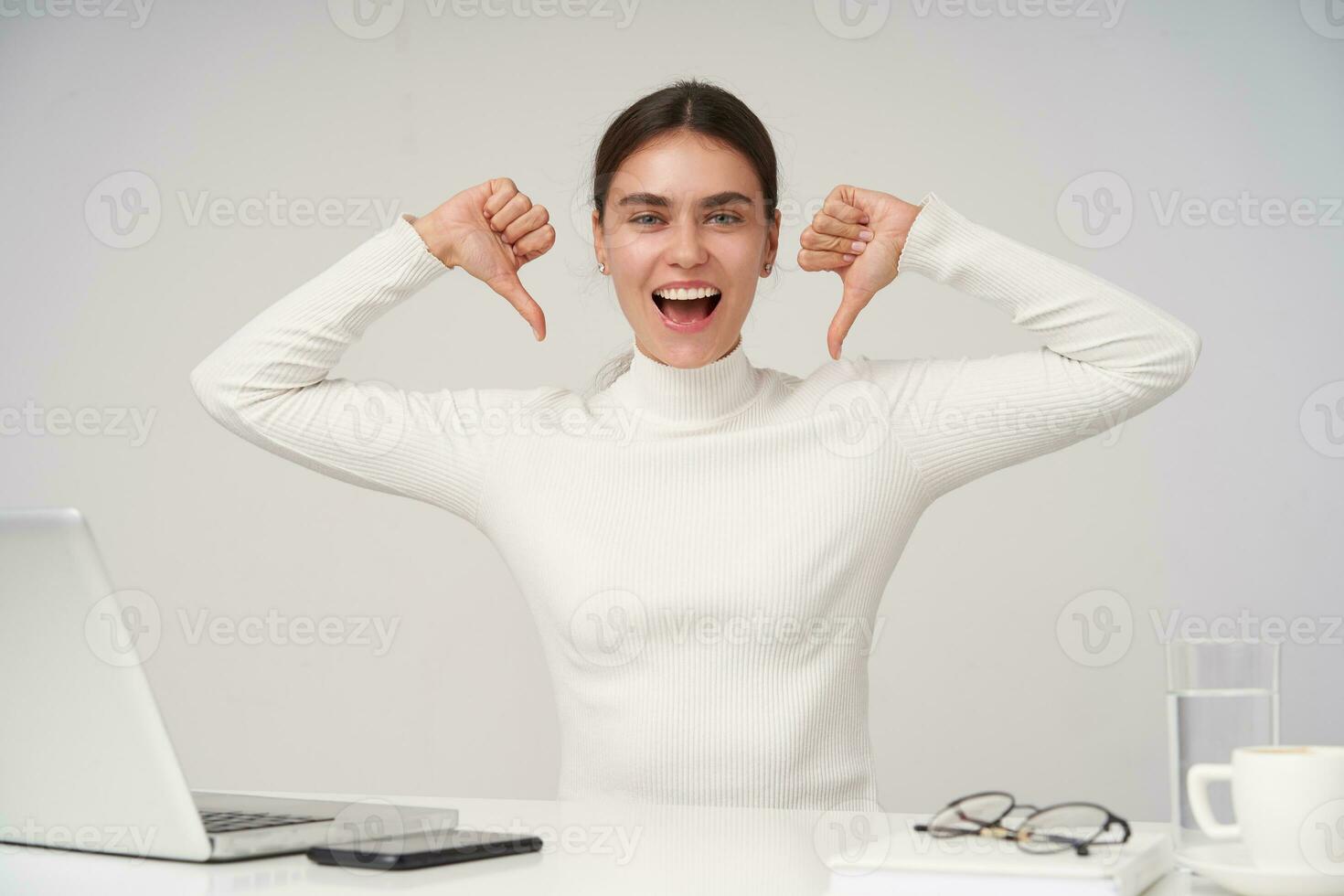 Joyful young attractive brunette female with natural makeup pointing happily to herself with raised hands and looking at camera with excited face, sitting at table over white background photo