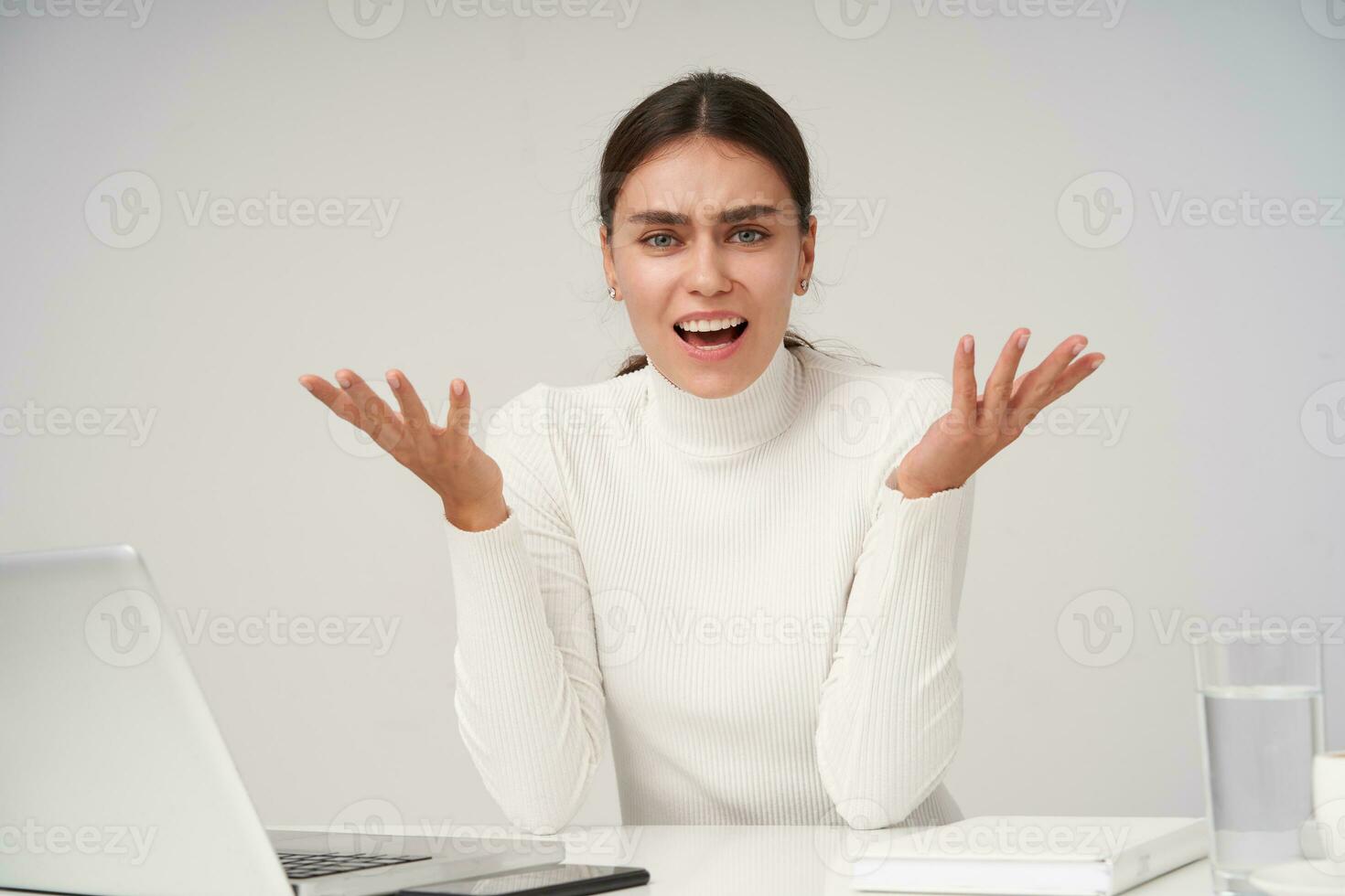Irritated young pretty dark haired female with natural makeup raising emotionally her hands while looking excitedly at camera, dressed in formal clothes while posing over white background photo