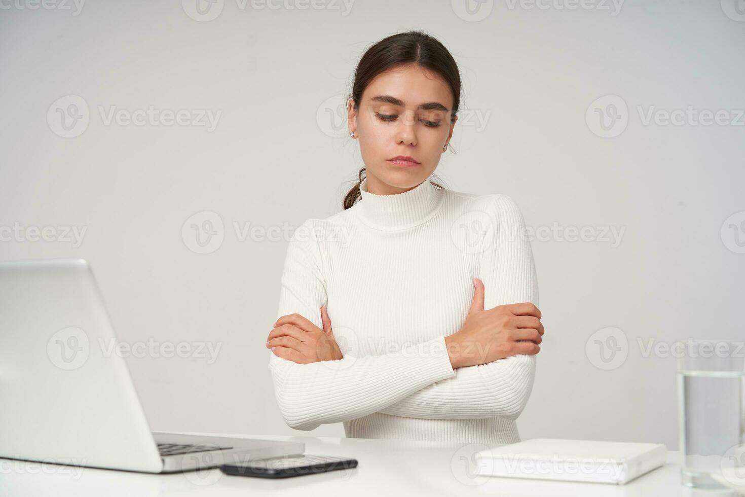 Upset young beautiful dark haired female with ponytail hairstyle folding hands on her chest and looking sadly down while sitting over white background with modern laptop photo