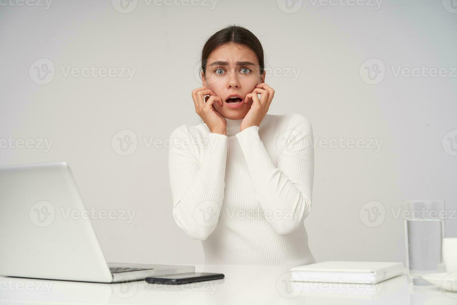 Frightened young dark haired woman in formal clothes holding her face with raised hands and looking amazedly at camera with wide eyes and mouth opened, posing over white background photo