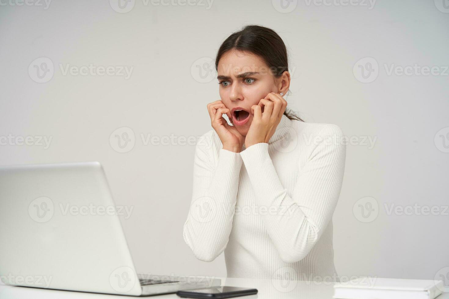 Shocked young pretty brunette woman with natural makeup keeping raised hands on her face while looking scaredly at screen of her laptop, isolated over white background photo