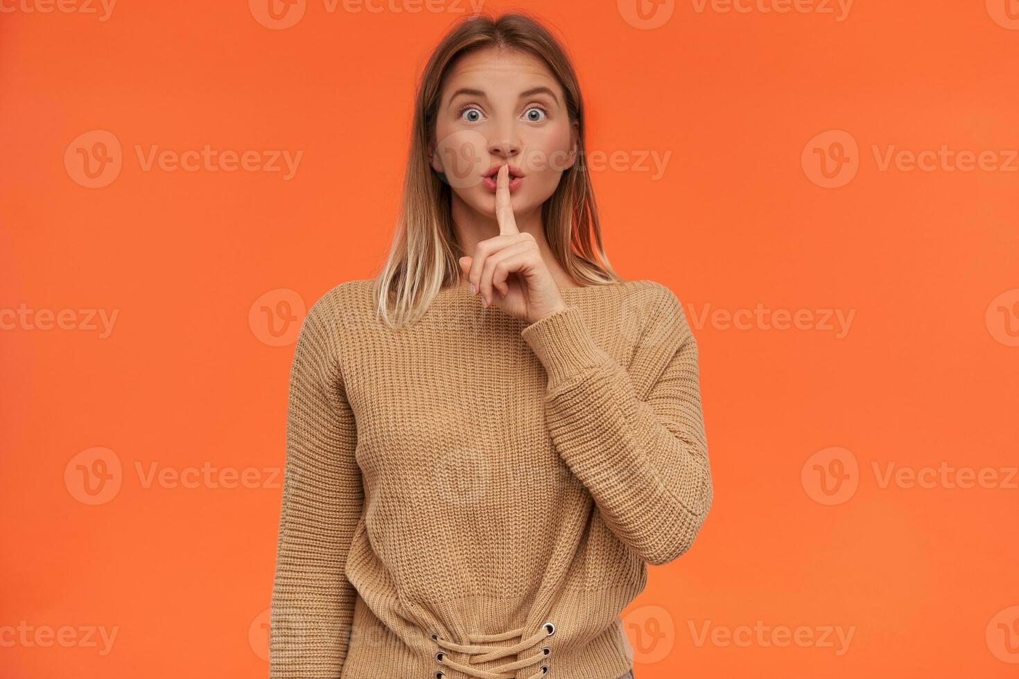 Excited young blue-eyed white-headed female raising hand in hush gesture to her mouth while looking emotionally at camera, standing over orange background photo