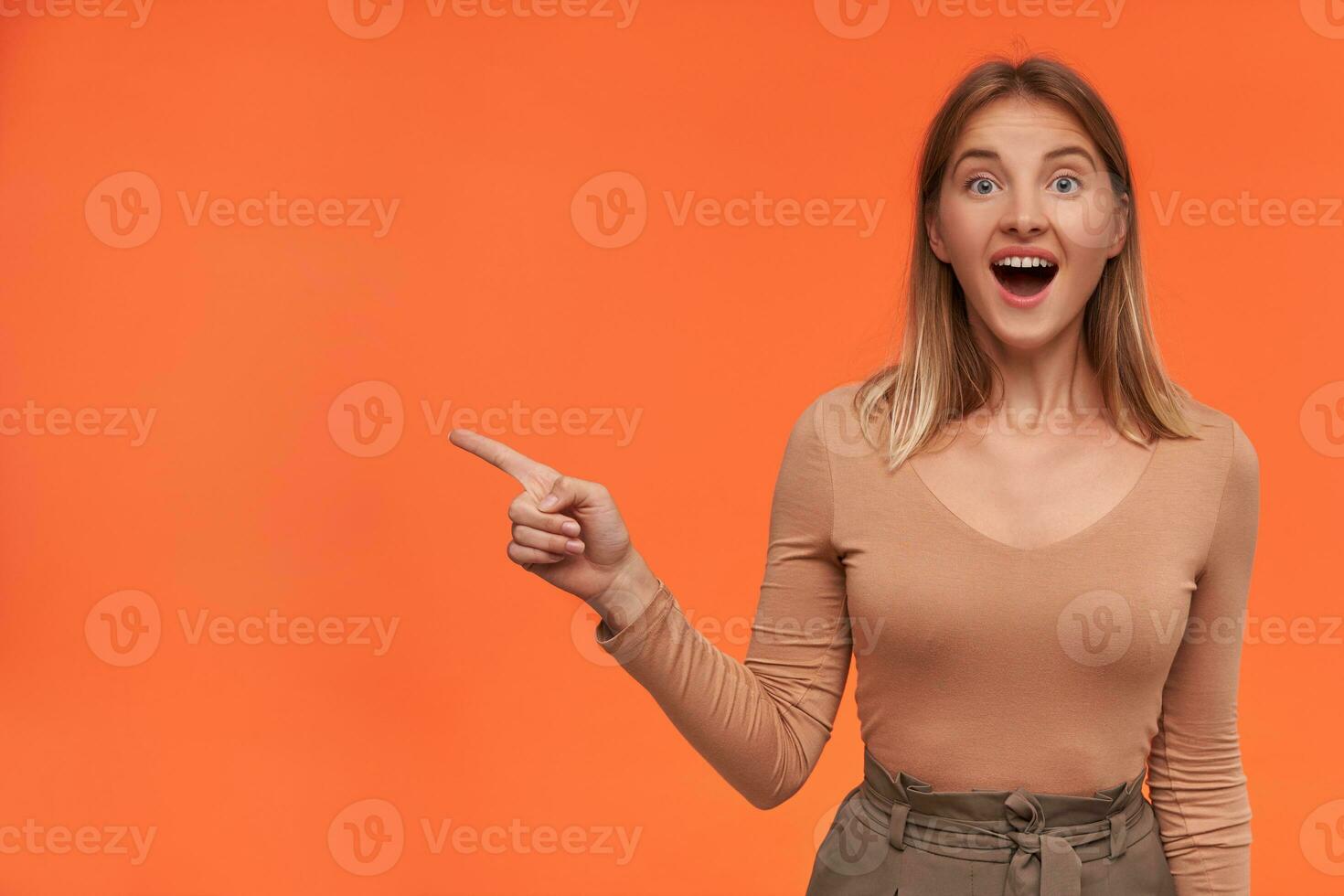 Excited young pretty blonde woman with short haircut looking amazedly at camera with wide eyes and mouth opened while showing aside, isolated over orange background photo