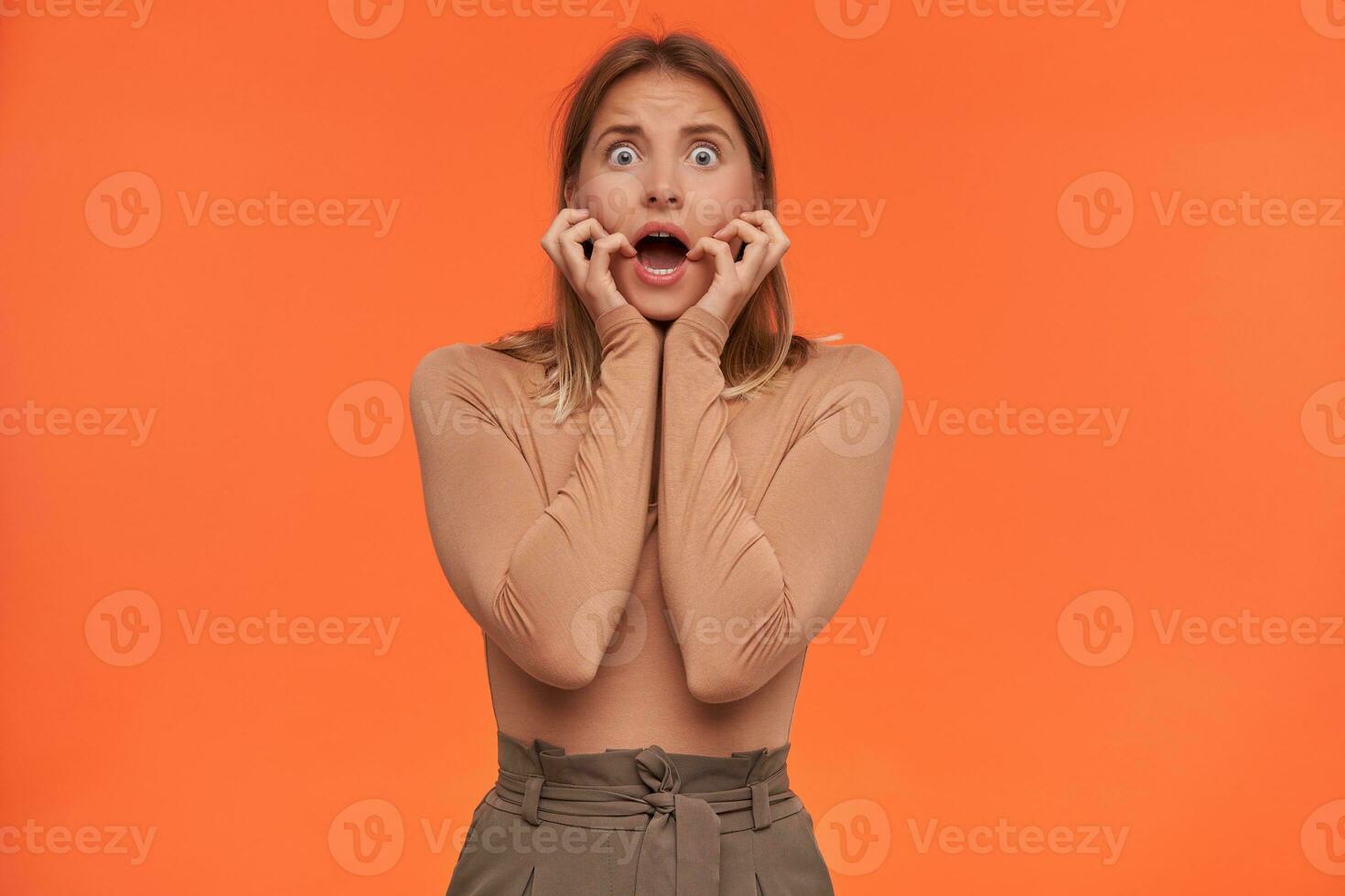 Open-eyed young pretty white-headed female with casual hairstyle keeping palms on her cheeks and grimacing face while looking scaredly at camera, posing over orange background photo