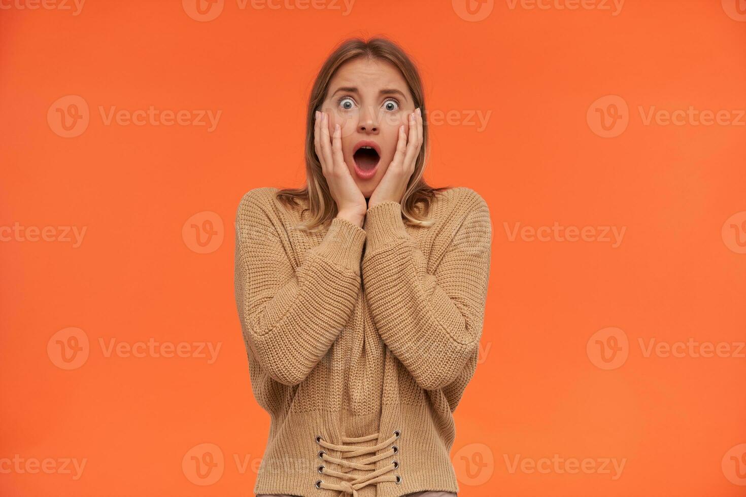 Shocked young pretty short haired white-headed woman with natural makeup holding her face with raised hands while looking dazedly at camera, isolated over orange background photo
