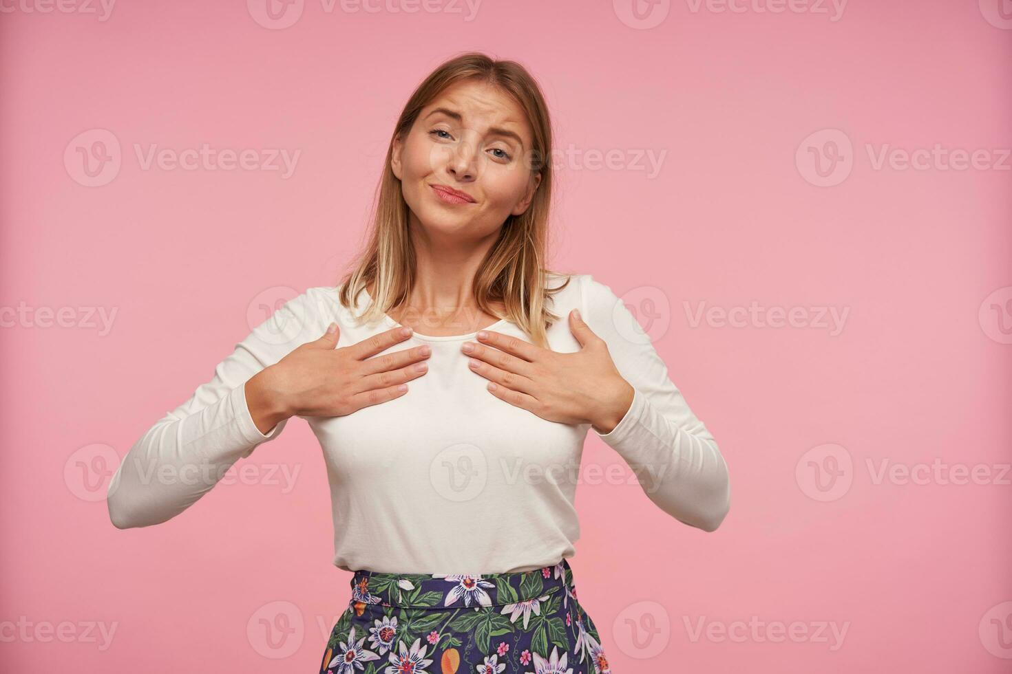 Indoor shot of positive lovely blonde lady in white blouse and flowered skirt looking at camera sincerely and holding raised palms on her chest, isolated over pink background photo